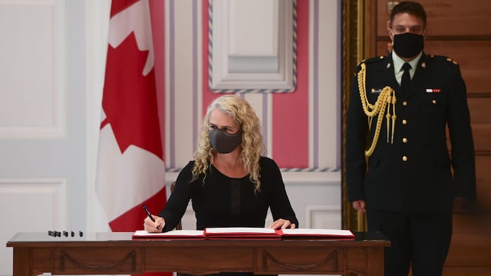 Elle est assise et signe un document, portant un couvre-visage, devant un drapeau du Canada et un agent en uniforme.