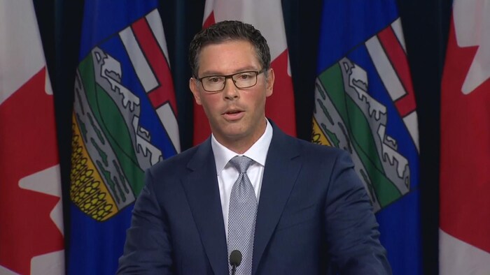 Un homme en costume cravate parle au podium devant une rangée de drapeaux de l'Alberta et du Canada.