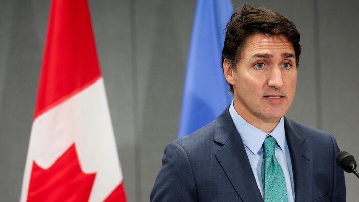 Justin Trudeau parle devant un micro. Derrière lui, il y a deux drapeaux.