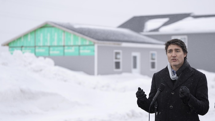 Justin Trudeau parle au microphone devant un monticule de neige et une bâtisse en construction.