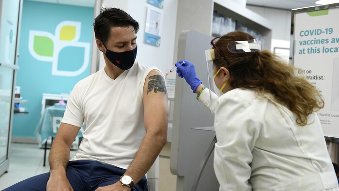Justin Trudeau, la manche de son t-shirt relevée, se fait vacciner par une femme. Les deux portent un masque.
