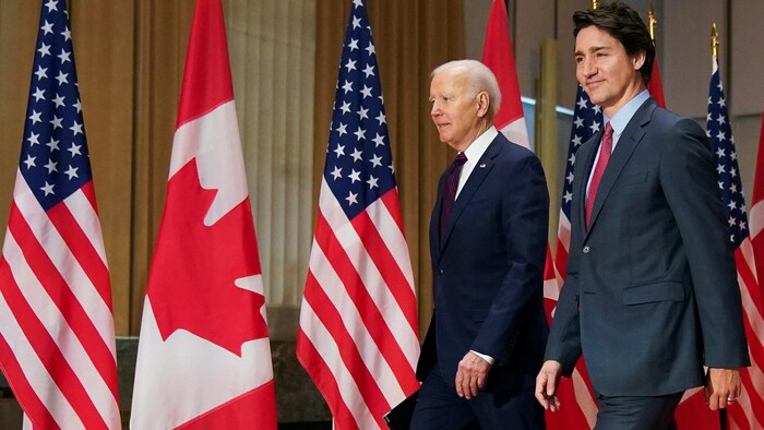 Joe Biden et Justin Trudeau marchent devant une rangée de drapeaux canadiens et américains.