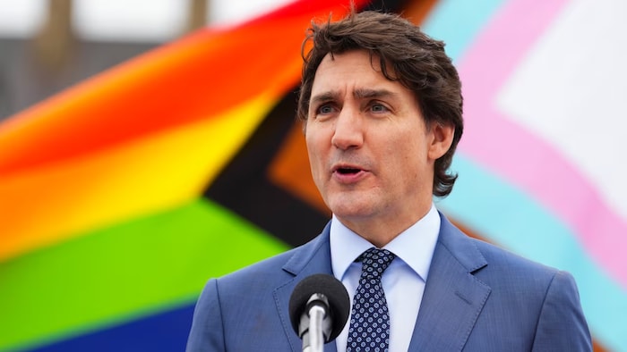 Justin Trudeau sa harap ng mikropono at sa background niya lumilipad ang Pride flag.