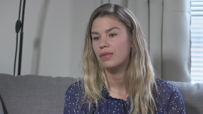 Kaitlin Bick est assise sur un canapé, elle a les cheveux blonds et longs, les yeux verts, elle est peu maquillée, elle porte un chandail violet avec des petits pois blancs. Elle regarde dans le vide.