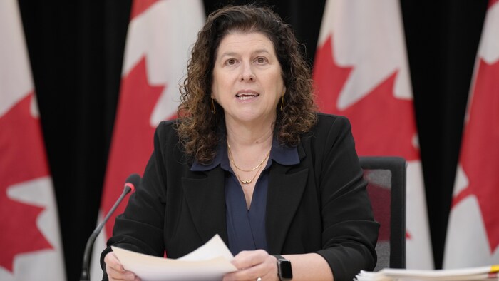 Une femme parle, feuilles en main, devant des drapeaux du Canada.