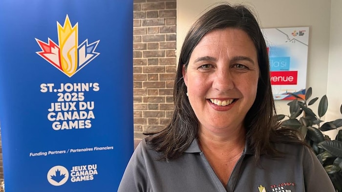 La PDG des Jeux du Canada 2025 pose devant le logo des Jeux qui représente la géologie accidentée du littoral terre-neuvien, l'architecture de Saint-Jean de Terre-Neuve et la flamme de la compétition sportive.
