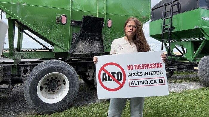 Une femme tient une pancarte signifiant qu'Alto n'a pas accès au terrain, devant de l'équipement agricole.