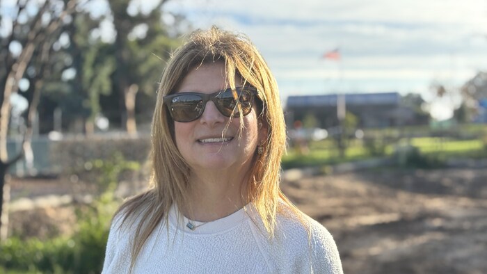 Karyn Weber pose avec des lunettes de soleil devant un terrain vide où flotte, au loin, un drapeau des États-Unis devant un bâtiment. 