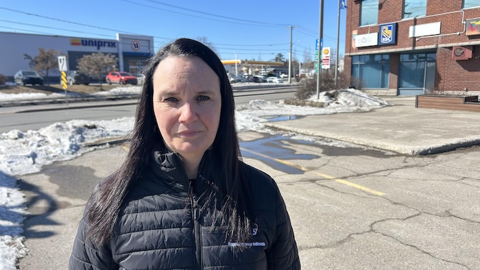 Une femme aux cheveux sombres, vêtue d'un manteau noir, se tient devant un stationnement fissuré avec de la neige fondante. En arrière-plan, on aperçoit un Uniprix, une station Shell et un bâtiment de briques rouges sous un ciel clair.
