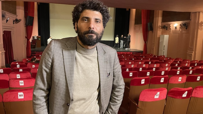 Un homme aux cheveux frisés et portant une petite barbe pose dans une salle de théâtre, le dos à la scène.