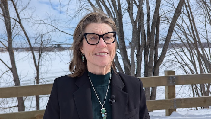 Une femme élégante, portant des lunettes en yeux de chat, sourit, debout en hiver le dos tourné au fleuve gelé et à une clôture en bois.
