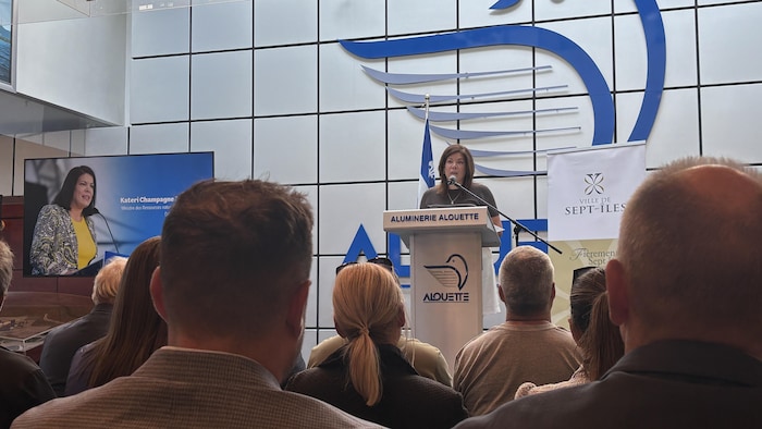 Vue de dos d'un public écoutant la ministre Kateri Champagne Jourdain parler derrière un lutrin arborant le logo de l'Aluminerie Alouette.