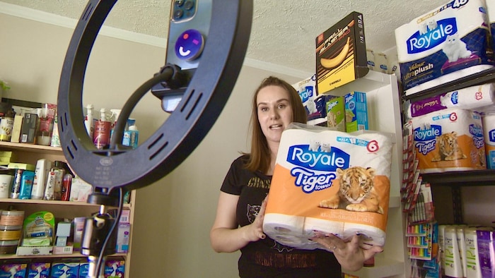 Kathleen Cassidy enregistre une vidéo pour ses réseaux sociaux devant une étagère de produits. Elle tient un paquet de papier toilette dans ses mains.