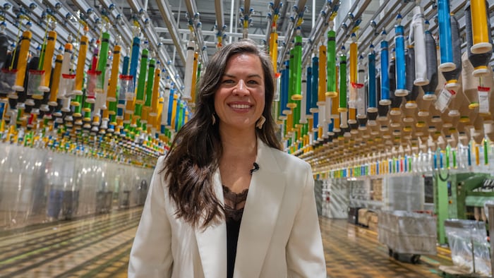 Une femme souriante sous des bobines de fil dans une usine de textile.
