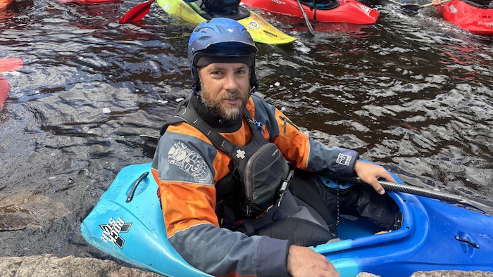 Un kayakiste barbu portant un casque bleu et une combinaison étanche est assis dans son kayak. Il se trouve sur l'eau, près du bord rocheux, avec d'autres embarcations visibles en arrière-plan.
