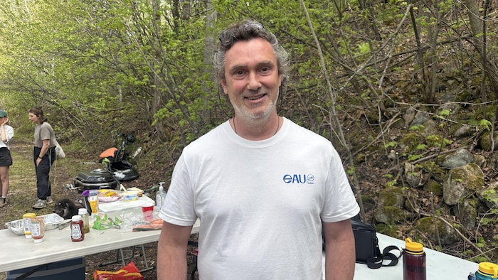 Un homme souriant en t-shirt blanc se tient au premier plan devant une table pliante de pique-nique. En arrière-plan, dans un décor de forêt, deux femmes discutent.