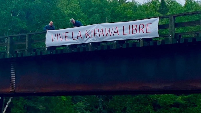 Deux personnes sur un pont au-dessus de la rivière Kipawa, tenant une banderole où il est écrit « Vive la Kipawa Libre ».