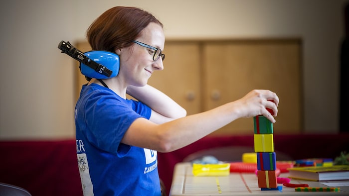 Une jeune femme rousse et souriante, photographiée de profil, tient d'une main des cubes multicolores empilés et de l'autre main, un casque bleu.