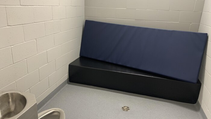 Une cellule provisoire, avec une toilette et un lavabo en inox ainsi qu'un lit simple.