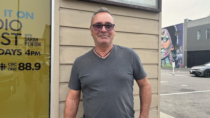 Un homme aux cheveux gris courts, portant des lunettes teintées et un chandail gris, se tient debout devant un mur de bois beige. Derrière lui, à droite, un stationnement et une murale colorée sur un mur d'immeuble. Le ciel est blanchâtre.