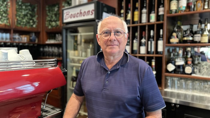 Un homme âgé aux cheveux blancs, portant des lunettes rondes et un polo bleu, se tient derrière le comptoir d'un restaurant.