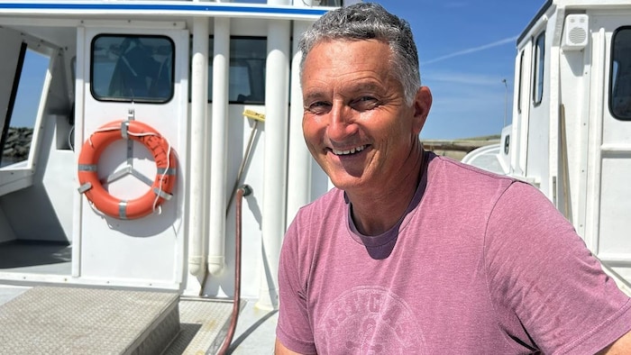 Un homme de belle apparence, aux cheveux argentés et au teint bronzé, est assis dans un rai de soleil sur le pont d'un bateau et sourit d'un air de connivence.