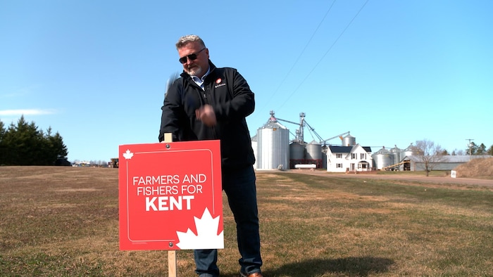 Kent Macdonald has a hammer in hand. He drives an electoral sign into the ground.