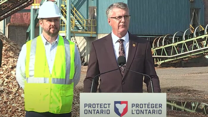 Le ministre se tient derrière un lutrin pour faire sa déclaration. Il est sur un chantier. Derrière lui se trouve un ouvrier vêtu d'une veste de sécurité et d'un casque.