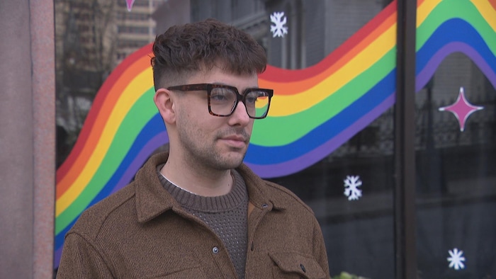 Un homme portant des lunettes à grosses montures noires et brunes est debout de profil devant une vitrine décorée d'un arc-en-ciel.