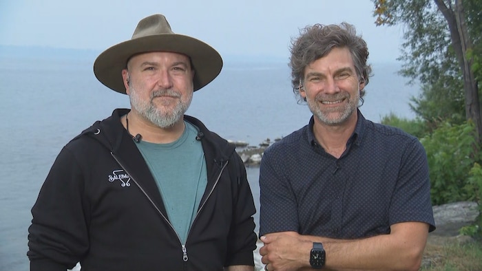 Deux hommes souriant devant un point d'eau et des arbres. 