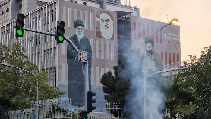 De la fumée s'élève dans une rue le long d'un immeuble sur lequel figure notamment la photo d'Ali Khamenei et d'autres personnages religieux iraniens.