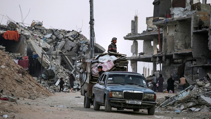 Des réfugiés sur une camionnette qui circule entre des édifices en ruine.