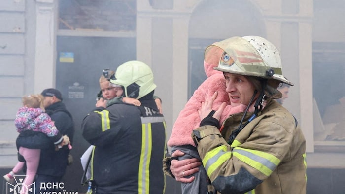 Trois pompiers marchent dans une rue en serrant dans leurs bras trois jeunes enfants qu'ils évacuent après une attaque de drone.