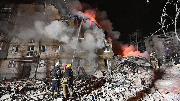 Des pompiers arrosent les flammes d'un incendie dans un immeuble partiellement détruit.