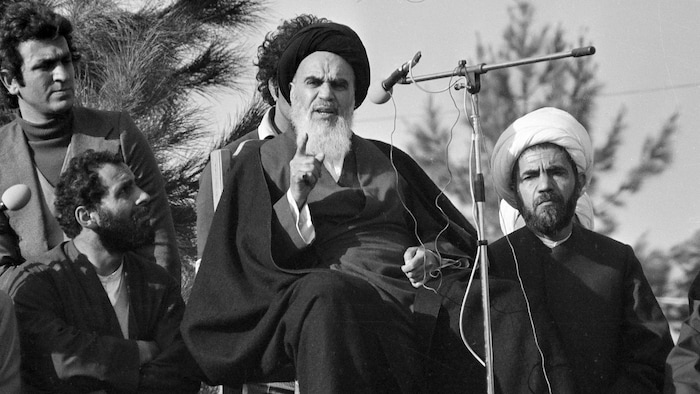L'ayatollah Khomeini parle devant un micro et il est entouré de quatre hommes.