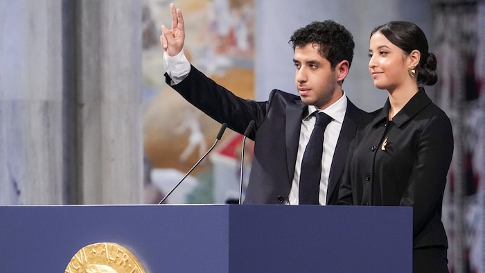 Une jeune femme et un jeune homme, qui fait le signe de la paix de sa main droite, sourient pensivement, debout derrière un lutrin.