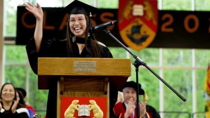 L'étudiante avec une toge et un chapeau de graduation sourit à une tribune, derrière un micro. Elle salue l'audience. 