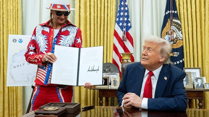 Un homme vêtu aux couleurs des États-Unis tient un décret signé et un homme est assis à un bureau.