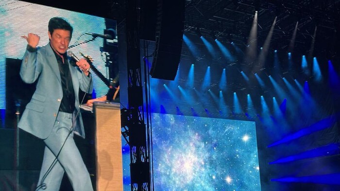 Le chanteur du groupe sur un écran géant, à côté de la scène qui a comme décor une galaxie. 