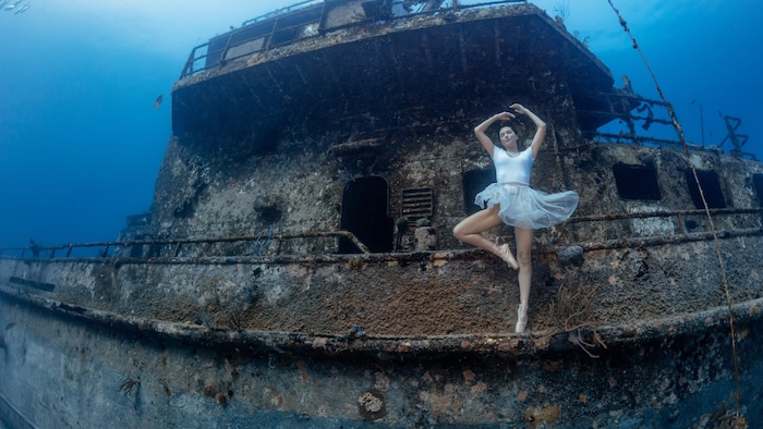 Une femme en tenue de ballerine sur une épave de bateau au fond de la mer. 