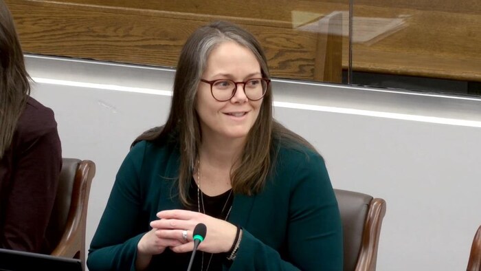 Kim Gill est assise à l'Assemblée législative, devant un micro. Elle tourne la tête vers la droite et sourit.