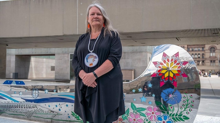 Kimberly Murray, portant un grand médaillon en perles autour du cou, se tient devant un grand canoë en acier inoxydable orné de motifs autochtones colorés.