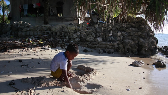 L'enfant joue dans le sable près de sa maison, construite à côté de l'eau et entourée d'un muret de pierre.