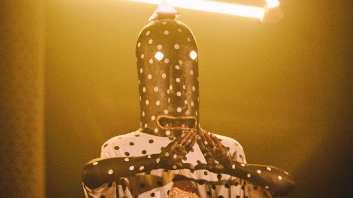 Le musicien costumé fait un signe de triangle avec ses mains, debout derrière sa batterie.