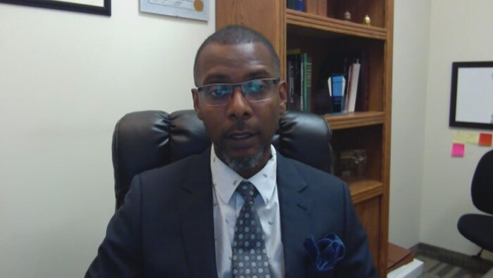 Un homme noir, portant des lunettes, vêtu d'un complet cravate, assis dans un bureau devant un écran.