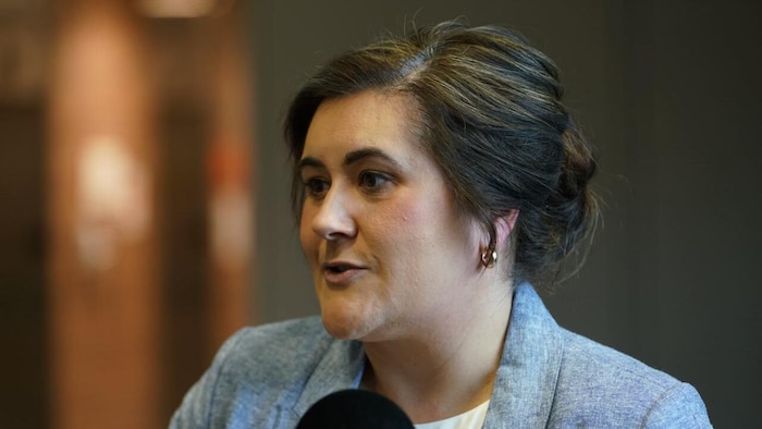 La ministre regarde vers sa droite et est en train de parler, avec plusieurs micros de différents médias pointés vers elle.