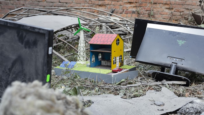 Une maquette représentant une maison, une éolienne et des panneaux solaires est intacte, au milieu de décombres.