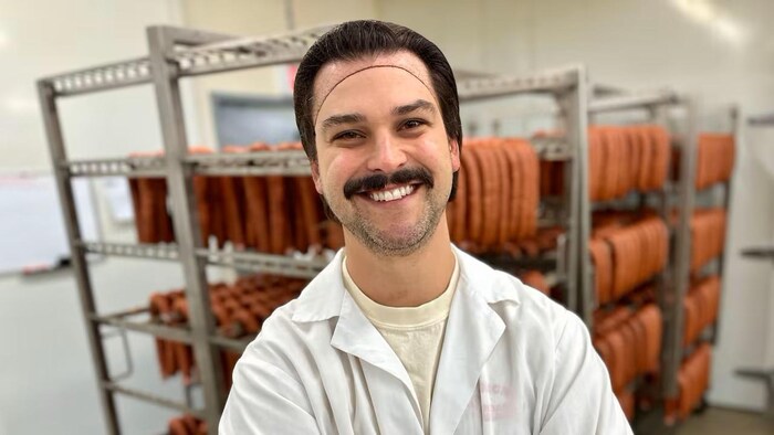 Kyler Zeleny sourit à la caméra. En arrière=plan, des saucisses produites par son usine.