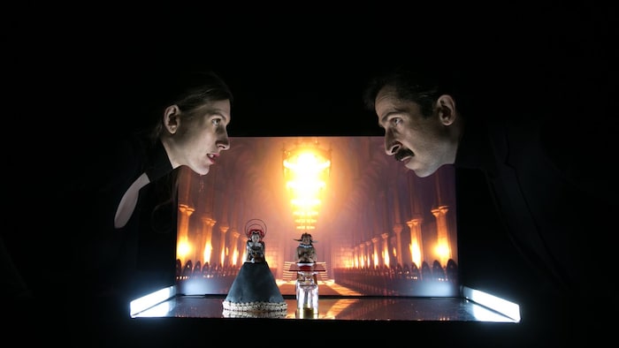 Un homme et une femme se regarde dans les yeux au dessus de deux poupées miniatures.
