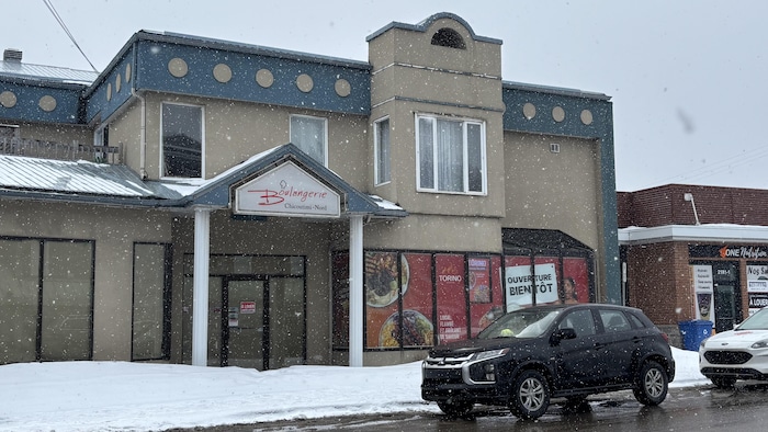 La photo montre la façade d'une boulangerie à Chicoutimi-Nord sous une chute de neige. Un véhicule noir est garé devant le bâtiment beige et bleu, tandis que de la neige accumulée recouvre le sol et le toit.